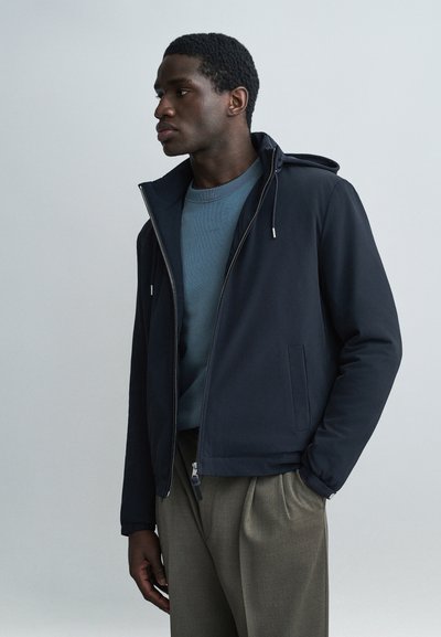 Jeune homme portant une veste zippée bleu marine par-dessus un sweat-shirt bleu et un pantalon beige, debout avec une main dans la poche, regardant vers la gauche sur un fond uni.