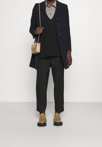 Marinblå ullkappa över en svart tröja med väst och rutigt skjorta, i kombination med svarta byxor och ljusbruna snörskor. Liten beige axelväska.