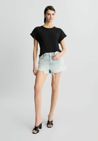 Černý top s krátkým rukávem v kombinaci s odrbanými světle modrými džínovými šortkami. Modelka má na sobě černé sandály na podpatku, minimální doplňky, neutrální pozadí.