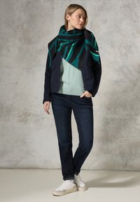 Lagdeling tøj med et grønt mønstret tørklæde, en navy jakke, en lys grøn sweater, mørke jeans og hvide sneakers med grå detaljer.