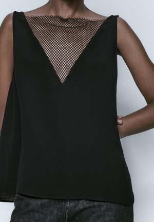 Ærmeløs sort top med dyb V-udskæring med et gennemsigtigt sort meshpanel, brugt med mørke bukser.