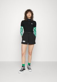 Top preto canelado de manga curta com gola de zip e mangas longas com padrão verde, combinado com calções pretos com um logótipo. Ténis pretos com solas brancas.