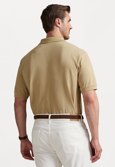 Polo bege de manga curta, com tecido texturizado e gola clássica, combinado com calças brancas e um cinto castanho.