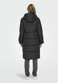 Long manteau en duvet noir avec capuche, comportant des sections matelassées horizontales et un design mi-long, associé à un pantalon gris et des chaussures légères.