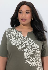 Frau mit langen dunklen Haaren, die goldene Creolen und ein olivgrünes Hemd mit weißen Blattmustern trägt und seitlich blickt.