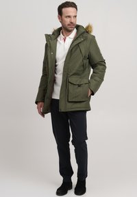 Parka acolchada de color verde oliva con capucha forrada de piel, que presenta múltiples bolsillos y un cierre de cremallera, llevada sobre pantalones oscuros y botas negras.