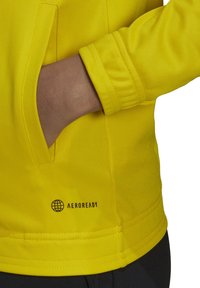 Žuta atletska jakna s glatkom, teksturiranom površinom, koja ima bočnu džep, rebraste manžetne i ispisani "AEROREADY" logotip blizu donjeg ruba.