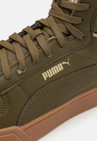 Olivově zelené tenisky s hladkým koženým svrškem, texturovanými detaily a hnědou gumovou podrážkou. Důrazná zlatá logo Puma.