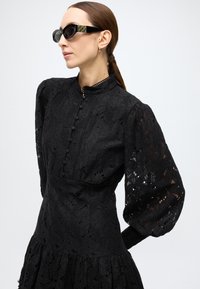 Black lace dress with puff sleeves, high neck, and button detailing. Worn with black sunglasses featuring a patterned side.