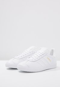 Vita lädersneakers med strukturerad gummisula, tre vita ränder och "GAZELLE"-logotyp i guld på sidan. Låg design.