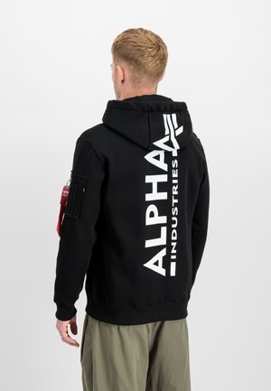 Mann mit kurzem blonden Haar, der einen schwarzen Alpha Industries Hoodie mit großem weißem Logo auf dem Rücken und olivgrüne Hosen trägt, steht seitlich.