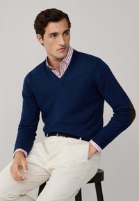 Suéter azul marino de cuello en V con una textura suave, que presenta parches marrones en los codos, usado sobre una camisa de dress a rayas rosas y pantalones beige.
