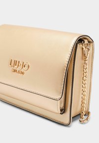 Kuldne metallik käekott sileda nahkpinnaga, mis sisaldab reljeefset logo, kettstrapi ja kontrastset õmblust ääredel.