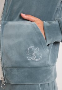 Roka rokā zilu pelēko velūru zipa jaku ar dekoratīvu rhinestone monogrammu "GL" uz priekšējā kabatas.