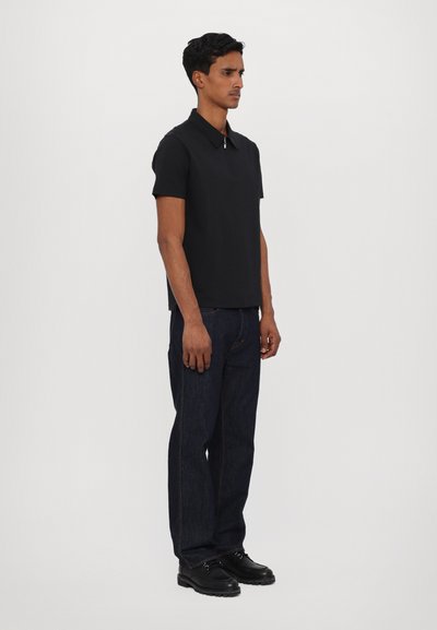 Camisa polo preta de manga curta combinada com jeans azul-escuro. A polo possui um colarinho clássico e um design ajustado. Sapatos pretos completam o visual.