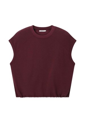 Femme en short bordeaux et haut sans manches, casquette verte, tenant un sac vert, portant des baskets marron avec des lacets roses, debout devant un fond beige.