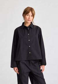 Černá košile na knoflíky s límečkem, dlouhými rukávy a volným střihem, s kontrastními knoflíky a hladkou texturou látky.