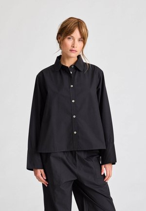 Must musta särk, millel on krae, pikad varrukad ja lõdvem lõige, kontrastsete nööpidega ja sileda kangaga.