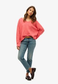 Jasnoróżowy, oversize sweter z szerokim dekoltem, zestawiony z jasnoniebieskimi dżinsami i burgundowymi mokasynami o teksturowanej podeszwie.