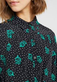 Blusa nera con pois bianchi e motivi floreali verdi. Presenta un colletto classico e una chiusura frontale con bottoni. Texture liscia.