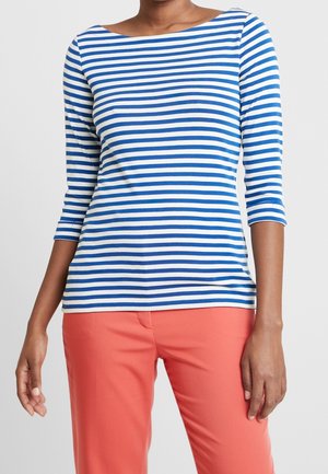 Kobieta ubrana w niebiesko-białą koszulę w poziome paski z długim rękawem, w połączeniu z koralowymi spodniami, na jednolitym tle.