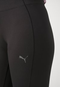 Mustad sportlikud retuusid venivast ja kehakujulisest kangast, vasakul reitel hall Puma logo ja lapikõmblused mugavuse tagamiseks.