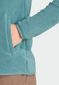 Pullover in pile di colore turchese, con una tasca laterale e una texture morbida. I polsini sono rifiniti e l'orlo inferiore è dritto.