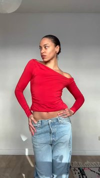 Γυναίκα με μαλλιά κολλημένα προς τα πίσω, φορώντας κόκκινο τοπ με μακριά μανίκια που αφήνει τους ώμους ακάλυπτους και ανοιχτόχρωμο μπλε τζιν, ποζάροντας με τα χέρια στη μέση σε φωτεινό δωμάτιο.