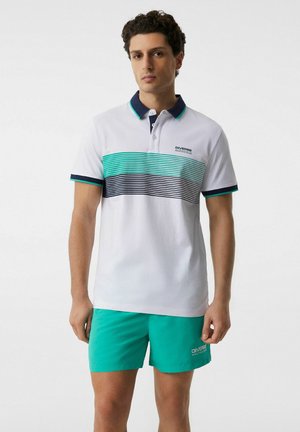 Młody mężczyzna w białej koszulce polo z turkusowymi i granatowymi paskami oraz turkusowych szortach, stojący na jednolitym tle.