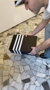 Black shoe box with three white stripes on the top side. The box is being lifted from a patterned stone floor.