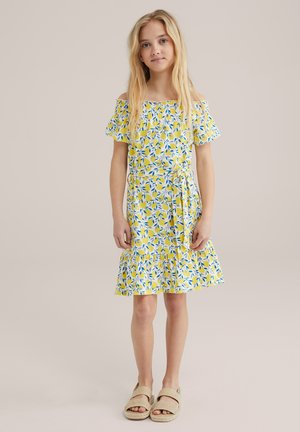 Gelbes Blumenkleid mit schulterfreien Ärmeln, gerafftem Saum und blauen Blättermustern. Trägt beige Riemchensandaletten.