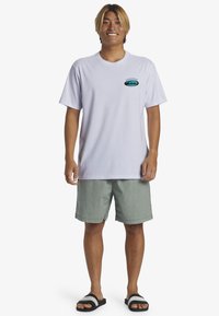 Camiseta blanca de algodón con mangas cortas y cuello redondo, que presenta un gráfico colorido en el pecho. Llevada con pantalones cortos verdes y sandalias negras.