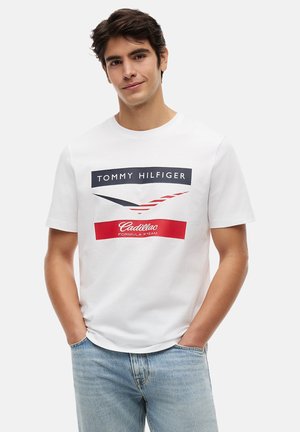Giovane uomo che indossa una maglietta bianca Tommy Hilfiger con il logo della squadra Cadillac Formula 1, abbinata a un paio di jeans azzurri, con le mani nelle tasche.