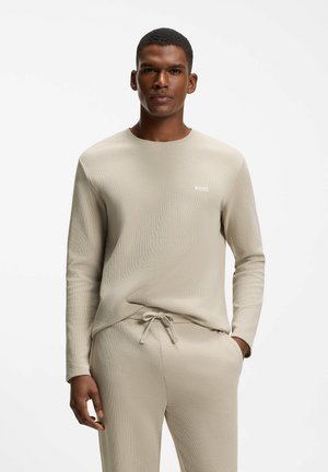 Man draagt een beige shirt met lange mouwen en bijpassende broek met trekkoord, staat met één hand in de zak tegen een witte achtergrond.