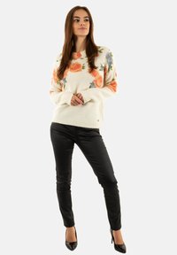 Mujer vistiendo un suéter blanco con patrones florales en naranja y azul, pantalones ajustados negros y zapatos de tacón alto negros, de pie con las manos entrelazadas.