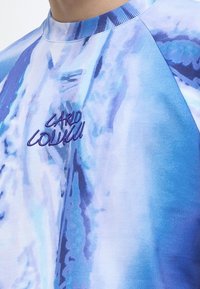Κοντινό πλάνο μπλε και λευκού t-shirt με batik, με το "Carlo Colucci" κεντημένο με σκούρο μπλε στο στήθος.