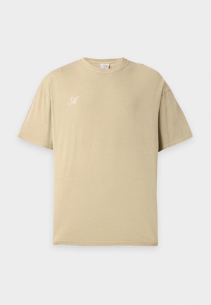 Mężczyzna z krótkimi włosami, ubrany w beżowy, oversize'owy t-shirt z białym napisem i ciemne spodnie, odwrócony tyłem na prostym jasnym tle.
