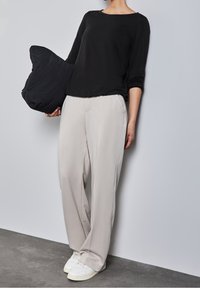 Svart långärmad topp, ljusgrå vida byxor och vita sneakers. Person som håller en svart kudde. Enkelt design med en avslappnad look.