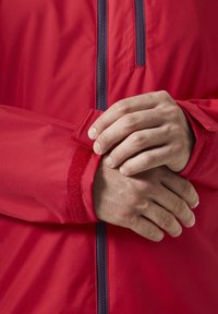 Czerwona wodoodporna kurtka z zamkiem błyskawicznym, regulowanymi mankietami na rzepy oraz gładkim, teksturowanym materiałem. Ręce umieszczone na mankietach.