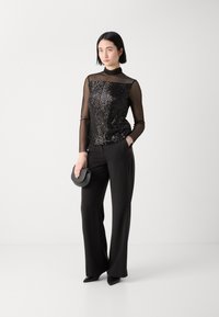 Schwarzes, mit Pailletten besetztes Oberteil mit transparenten langen Ärmeln, kombiniert mit weiten schwarzen Hosen. Das Model hält eine kleine schwarze Handtasche.
