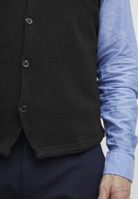 Svart stickad väst med fem framknappar, böjd nederkant, lagd över en ljusblå långärmad skjorta. Marinblå byxor delvis synliga.