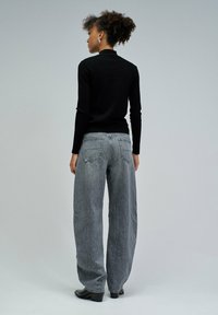 Kobieta z kręconymi włosami w czarnym golfie i szerokich, wyblakłych szarych dżinsach stoi tyłem na tle jasnoszarym.