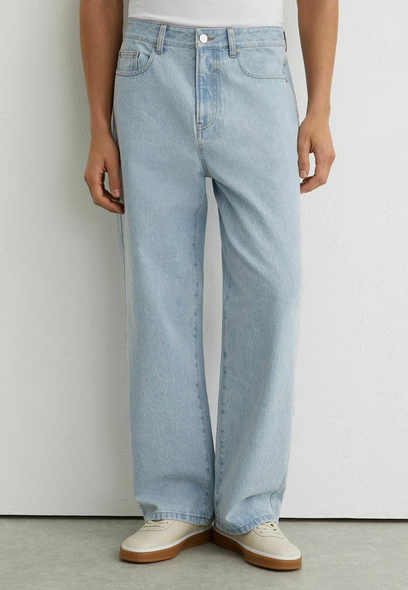 Personne portant un jean bleu clair à jambes larges et des baskets beiges, debout contre un mur blanc, les mains détendues le long du corps.