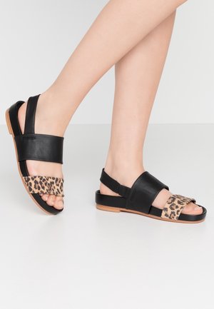 Pieds portant des sandales plates noires avec des brides imprimées léopard beige sur fond blanc uni.
