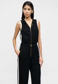 Μαύρη ολόσωμη φόρμα με κουκούλα, κλείσιμο με χρυσό φερμουάρ και ζωνάκι στη μέση. Διαθέτει κρεμ ρίγες στα πλάγια και άνετη γραμμή.