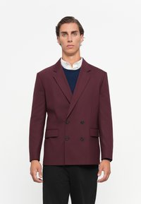 Bordeaux dobbeltradet blazer med sorte knapper, reverskrave, siderlommer og en skræddersyet pasform, lagd over en marineblå sweater.