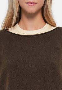 Sweater canelado castanho escuro com um decote largo, sobre uma t-shirt bege clara. Textura suave, design casual e detalhe de costura visível.