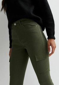 Pantalones cargo verde oliva hechos de una mezcla de algodón, con bolsillos frontales y un bolsillo lateral con solapa, combinados con un suéter de punto negro.