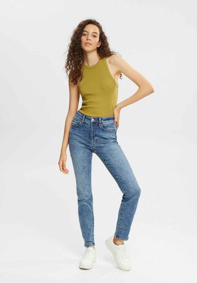 Olivgrünes ärmelloses Top mit geripptem Textur, kombiniert mit hochgeschnittenen hellblauen Jeans und weißen Turnschuhen. Einfaches und modernes Design.