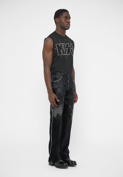 Camiseta gráfica negra sin mangas con letras con tachuelas, combinada con jeans oscuros con un detalle lateral brillante y zapatos negros.